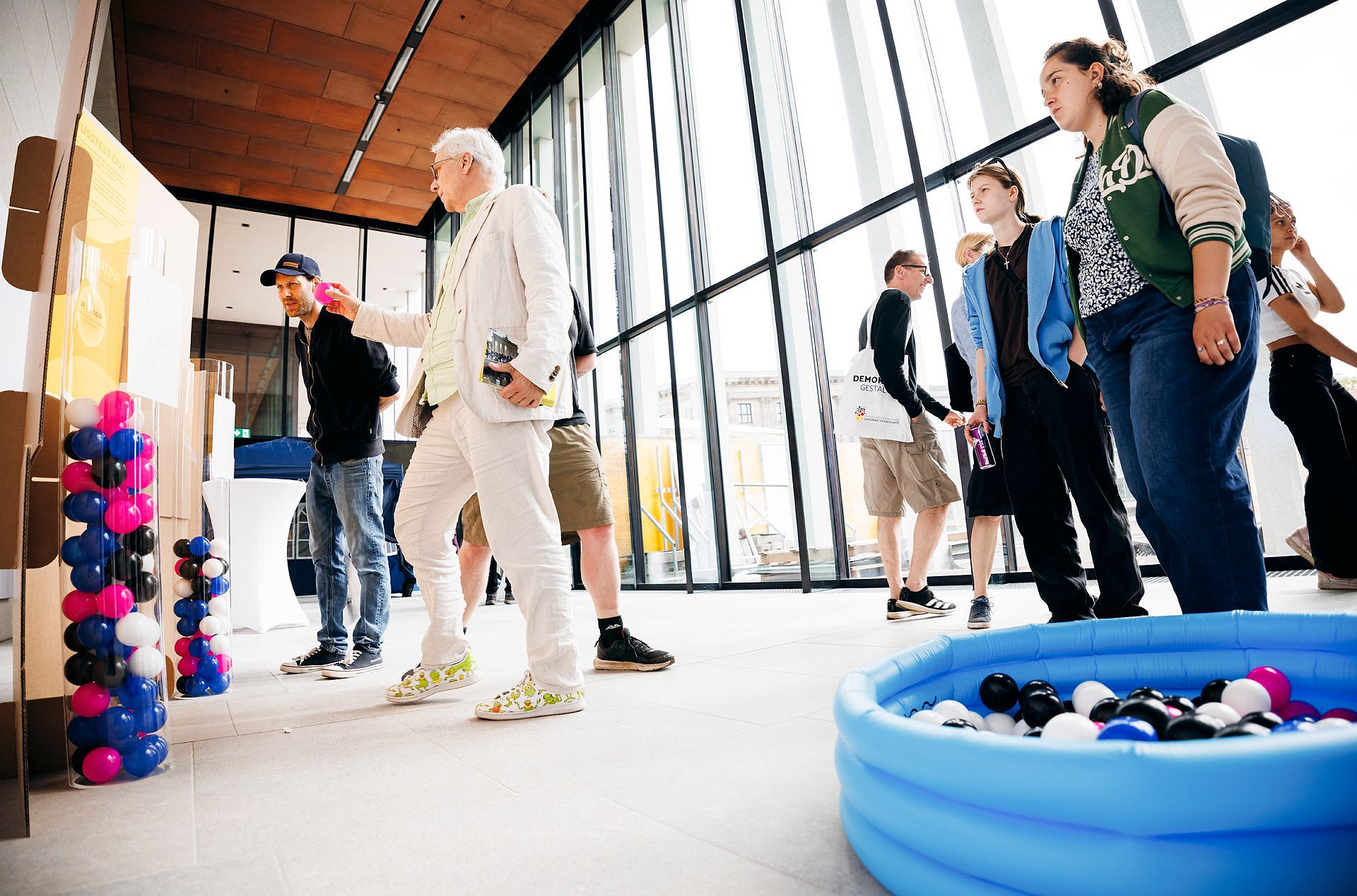Menschen werfen bunte Bälle aus einem Planschbecken in Plexiglassäulen