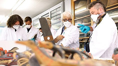 Mehrere Menschen in Kitteln und mit Masken in einem Depot an Objekten