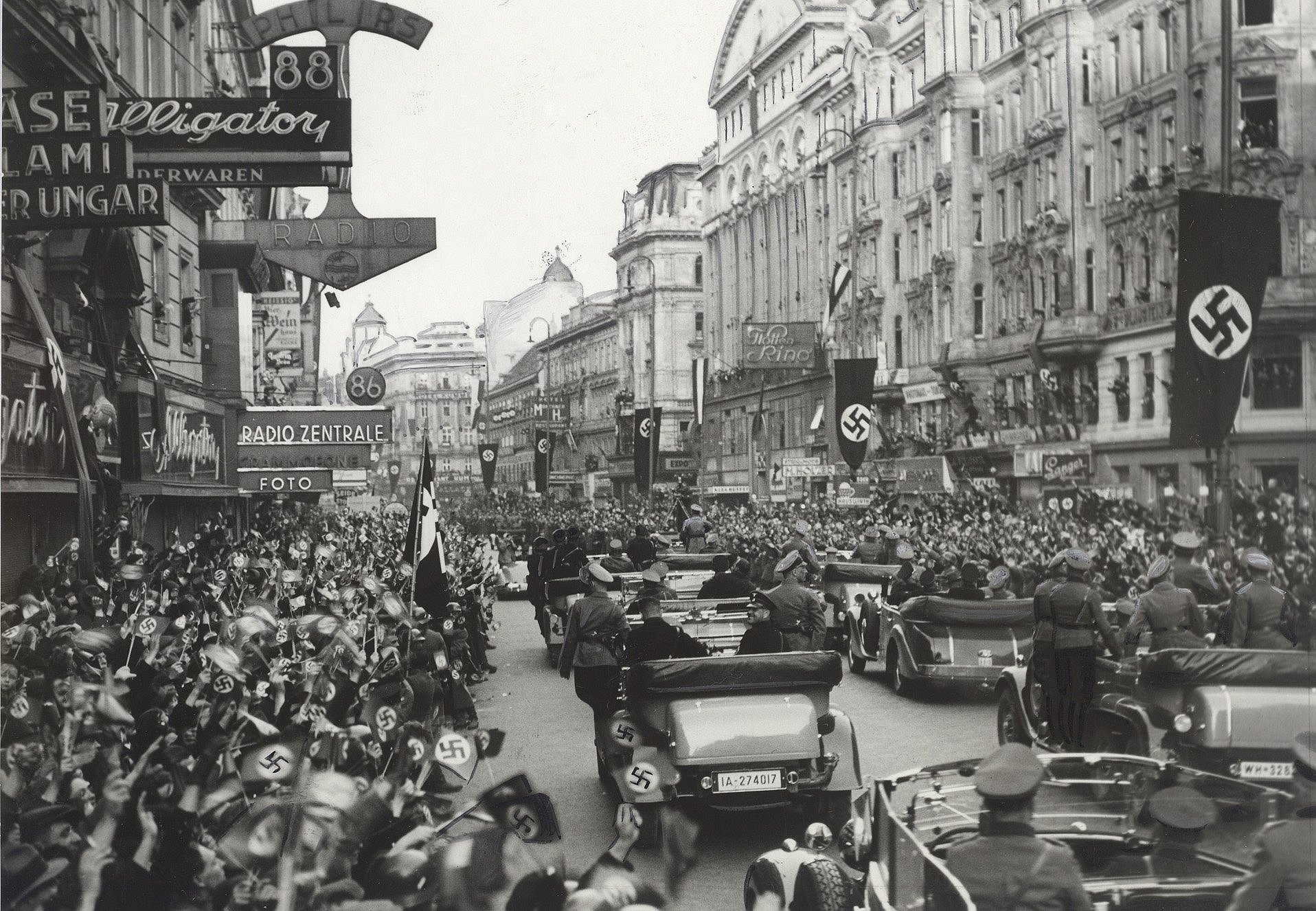 Historisches Foto einer Autokolonne in einer Straße voll Menschen und Hakenkreuzfahnen