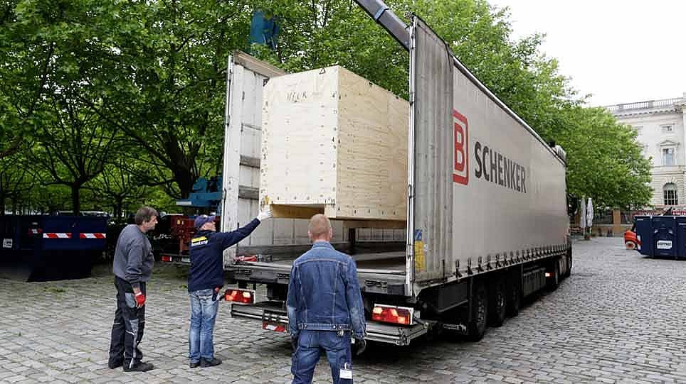 Eine große Holzkiste und mehrere Personen steht auf einem geplasterten Platz vor einem Fahrzeug (öffnet Vergrößerung des Bildes)