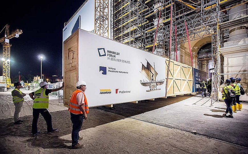 Mehrere Arbeiter mit Bauhelmen beobachten, wie bei Nacht eine große Holzkiste durch die Pforte eines eingerüsteten Gebäudes gebracht wird (öffnet Vergrößerung des Bildes)