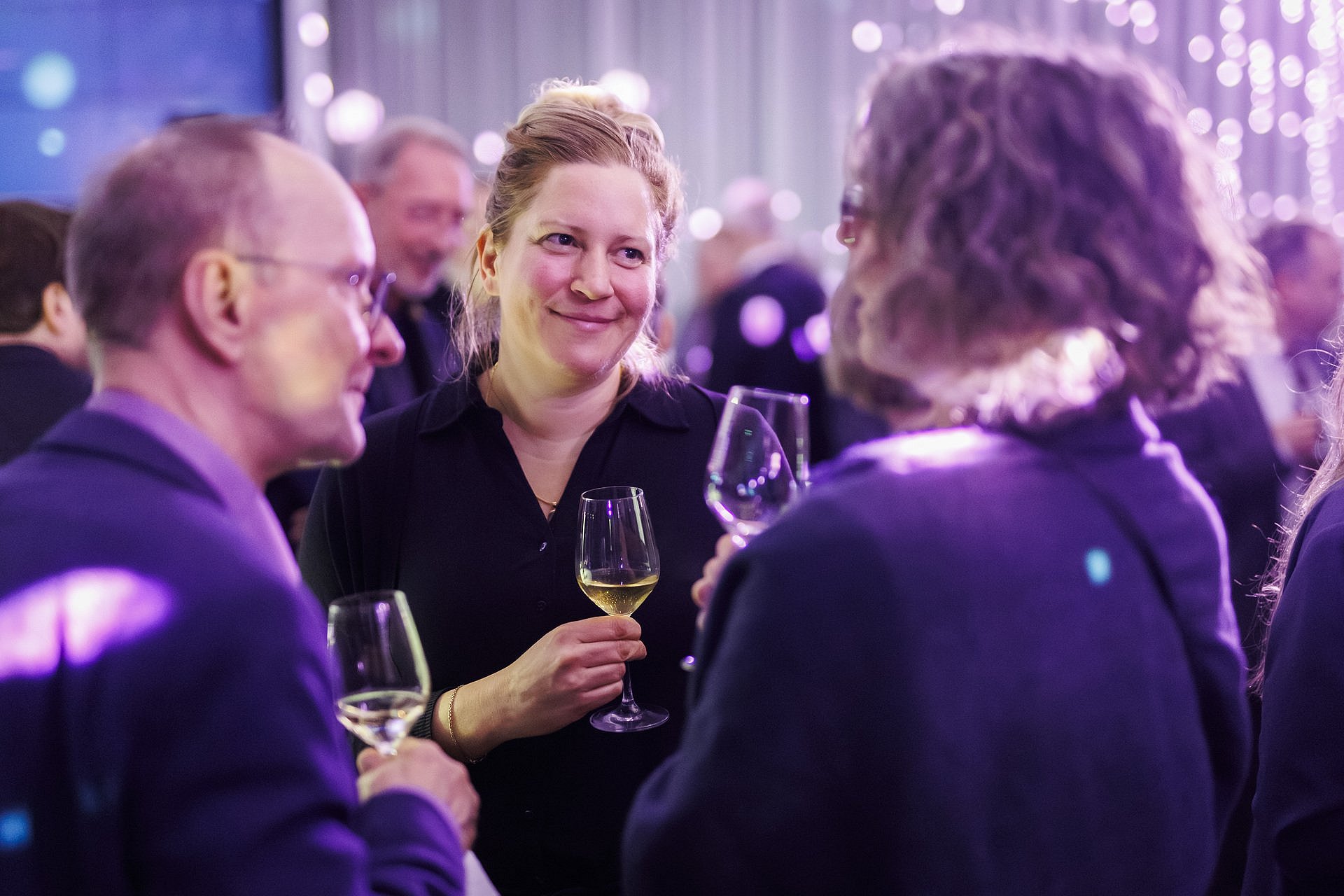 Drei Gäste stehen bei einem Empfang zusammen und unterhalten sich mit Weingläsern in der Hand; im Hintergrund weitere Gäste und stimmungsvolle Beleuchtung.