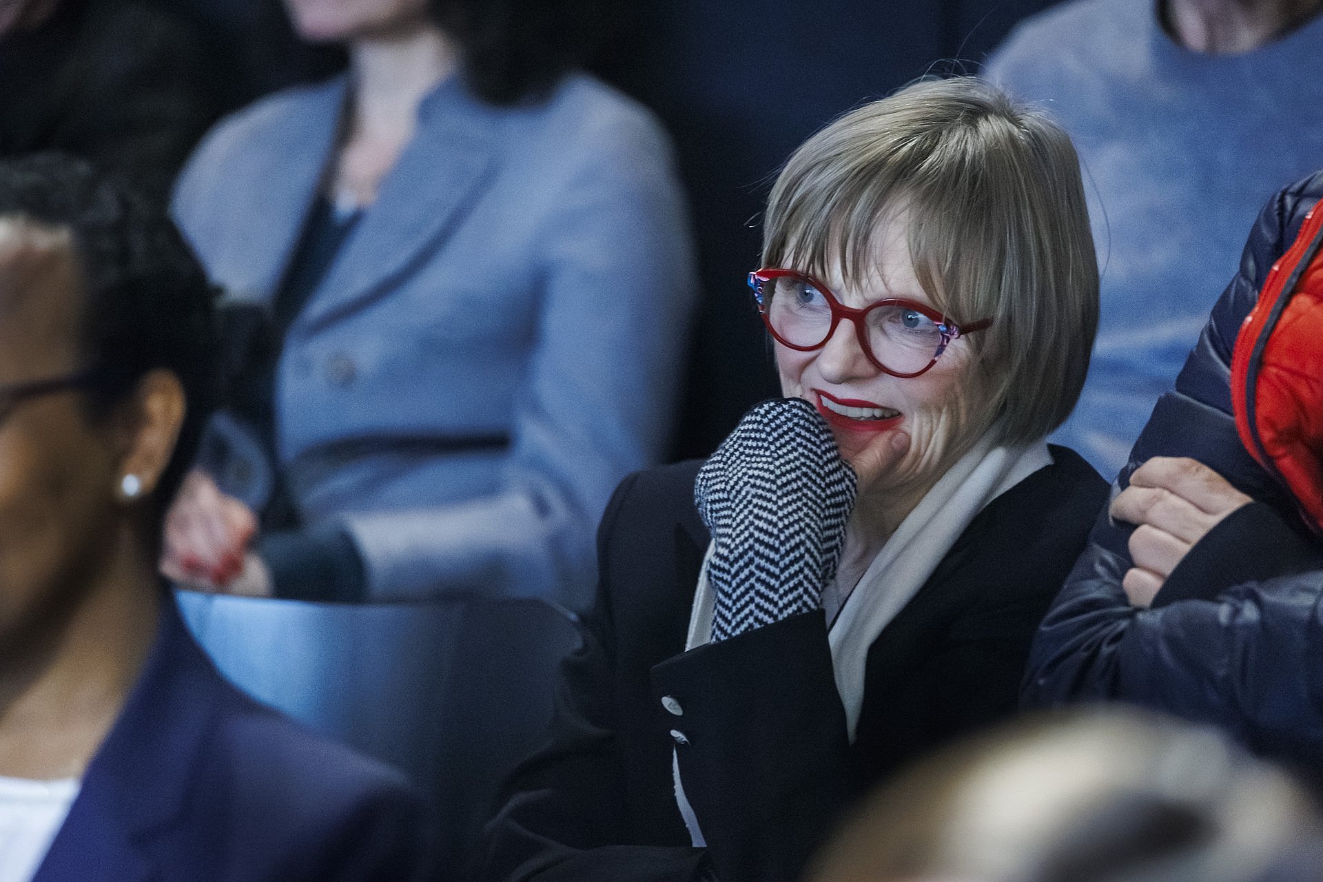 Eine Frau mit roter Brille sitzt im Publikum und lächelt, während sie dem Bühnenprogramm zuhört; weitere Gäste sind im Hintergrund zu sehen.
