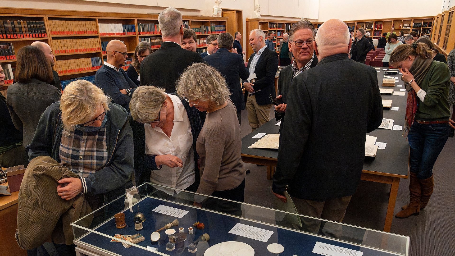 A group of people in a reading room. Some are leaning over their desks, others are chatting.