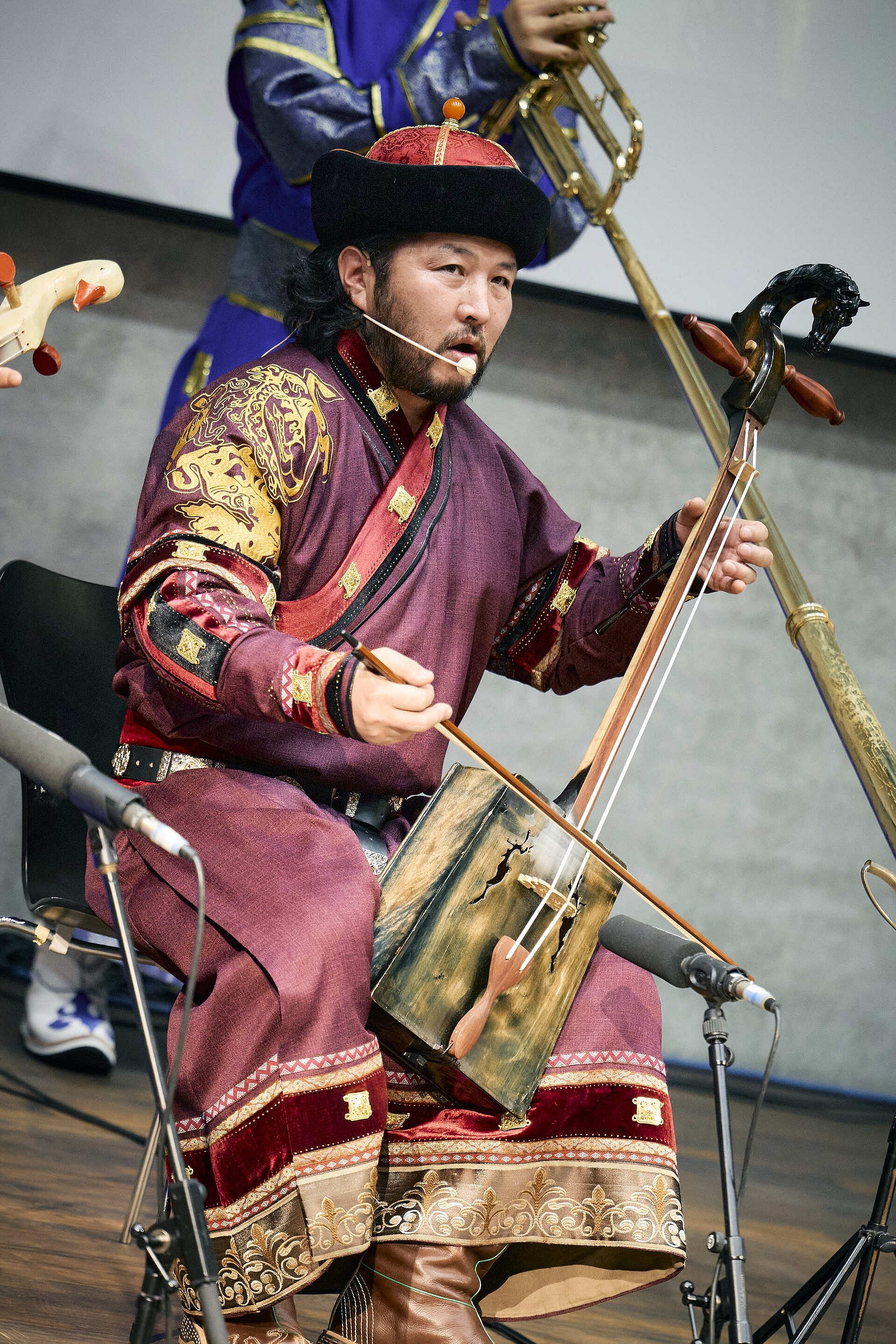 Ein sitzender Musiker in einem verzierten Gewand spielt ein Streichinstrument mit trapezförmigem Korpus und geschnitztem Pferdekopf.