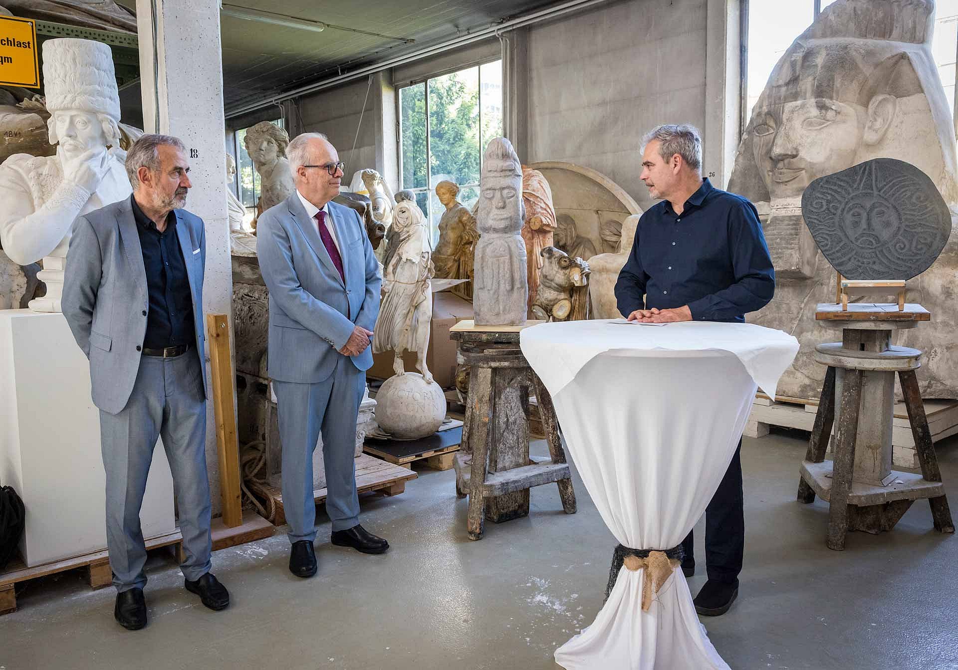 Zwei Männer hören einem anderen Mann zu, im Hintergrund Steinobjekte