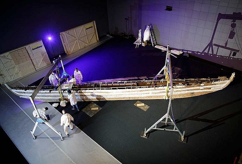 Blick von oben in einen musealen Innenraum, in dem ein hölzernes Boot verpackt und von mehreren Personen zum Transport vorbereitet wird (öffnet Vergrößerung des Bildes)
