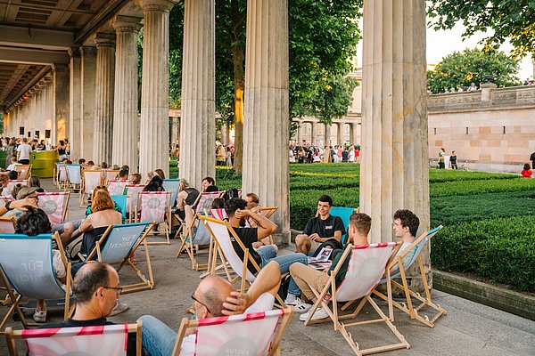 Junge Menschen sitzen in Sonnenstühlen unter einem Kolonnadengang, es ist Sommer
