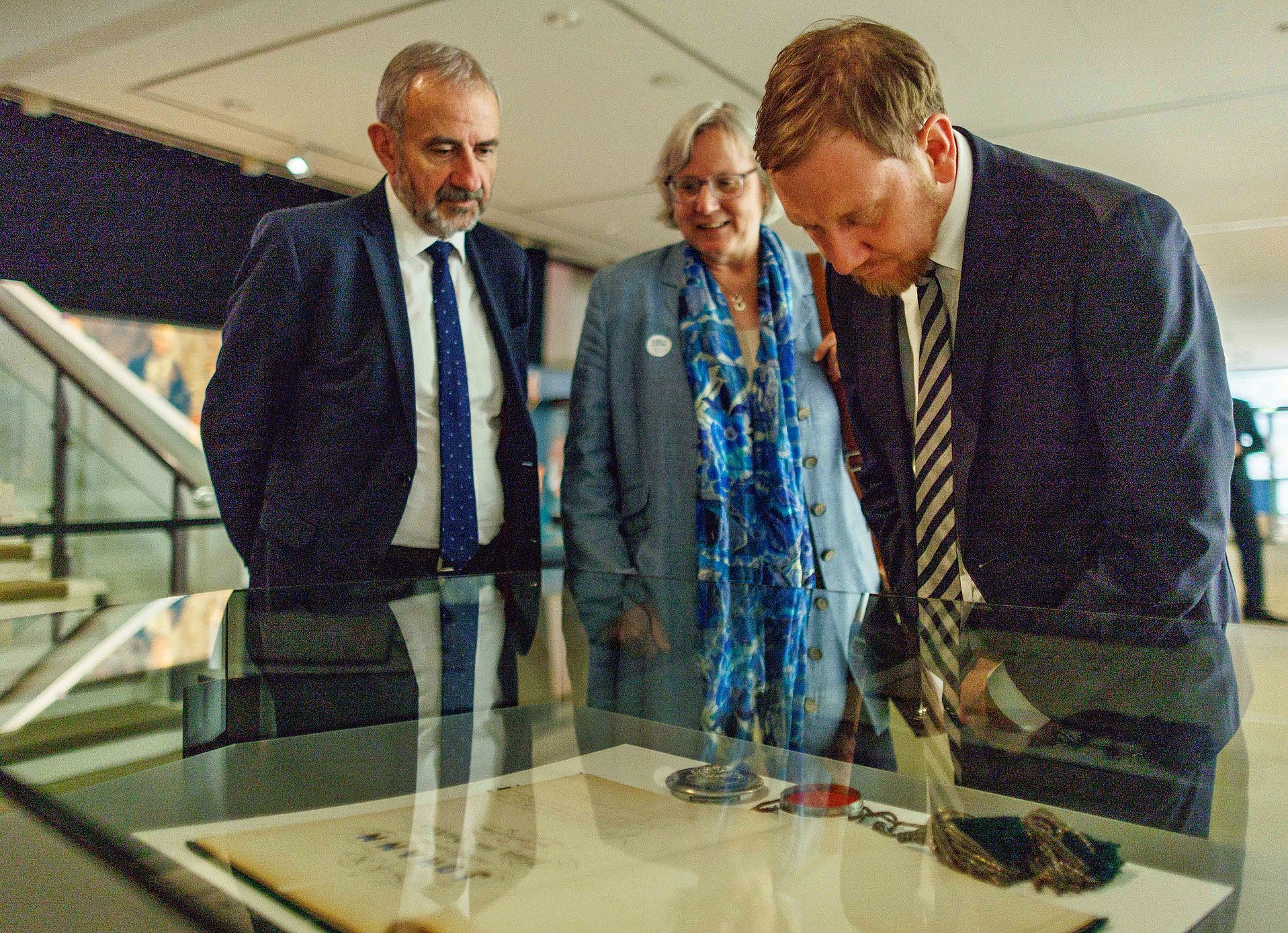Zwei Männer und eine Frau beugen sich über ein Schriftstück in einer Vitrine