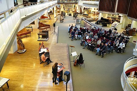 Menschen sitzen auf Stuhlreihen, auf einem Podest in einem Musikinstrumenten-Museum musizieren drei Personen.