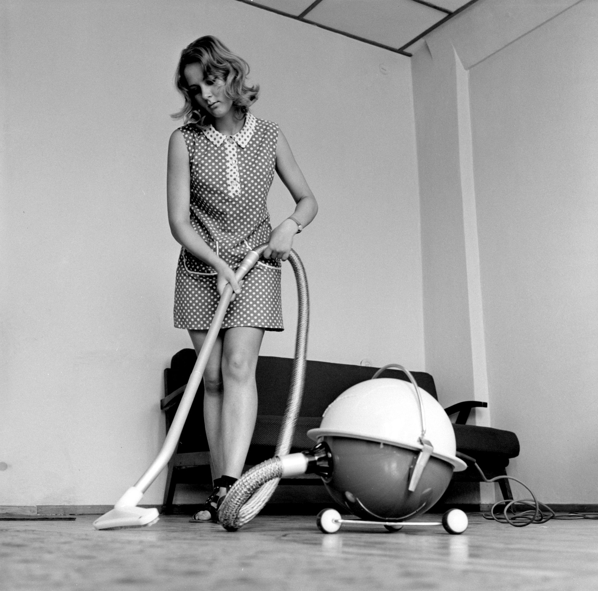 Schwarzweißfoto einer Frau in 60er-Jahre-Kleidung mit einem runden Staubsauger