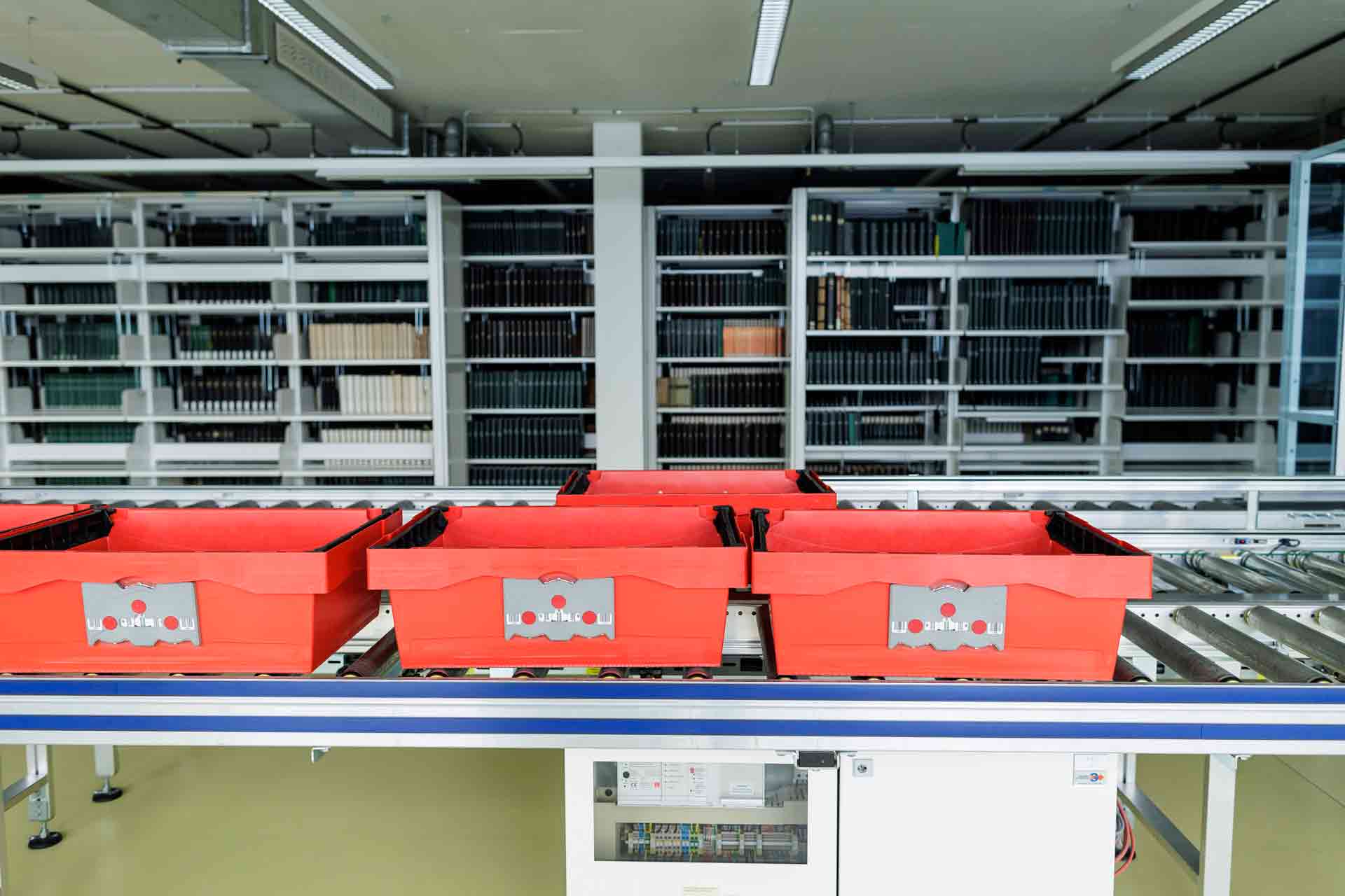 Rote Transportboxen auf einem Rollband, dahinter Archivregale